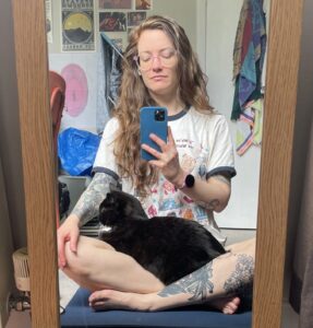 mirror selfie pic of a woman looking at her phone with a cat laying in her lap, on a yoga mat. In the background is white/beige walls and a door covered in some colorful posters and towels hanging as well as an IKEA blue bag.