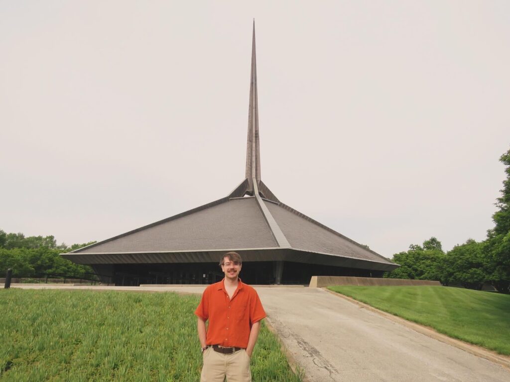 A photograph of a man with a orange shirt, short brown hair, and a moustache standing in the foreground. In the background, is the North Christian Church by Eero Saarinen, a pentagram-shaped building with a tall spire in the center.