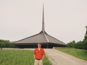A photograph of a man with a orange shirt, short brown hair, and a moustache standing in the foreground. In the background, is the North Christian Church by Eero Saarinen, a pentagram-shaped building with a tall spire in the center.