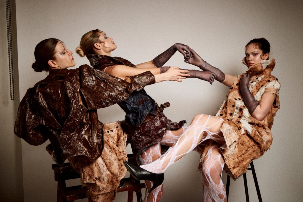 Three models wearing experimental fashion garments in brown, rust and pale textured fabrics pose together on chairs. The looks feature sculptural silhouettes, raw edges, buttons, crystallised surface details and white cut-out tights.