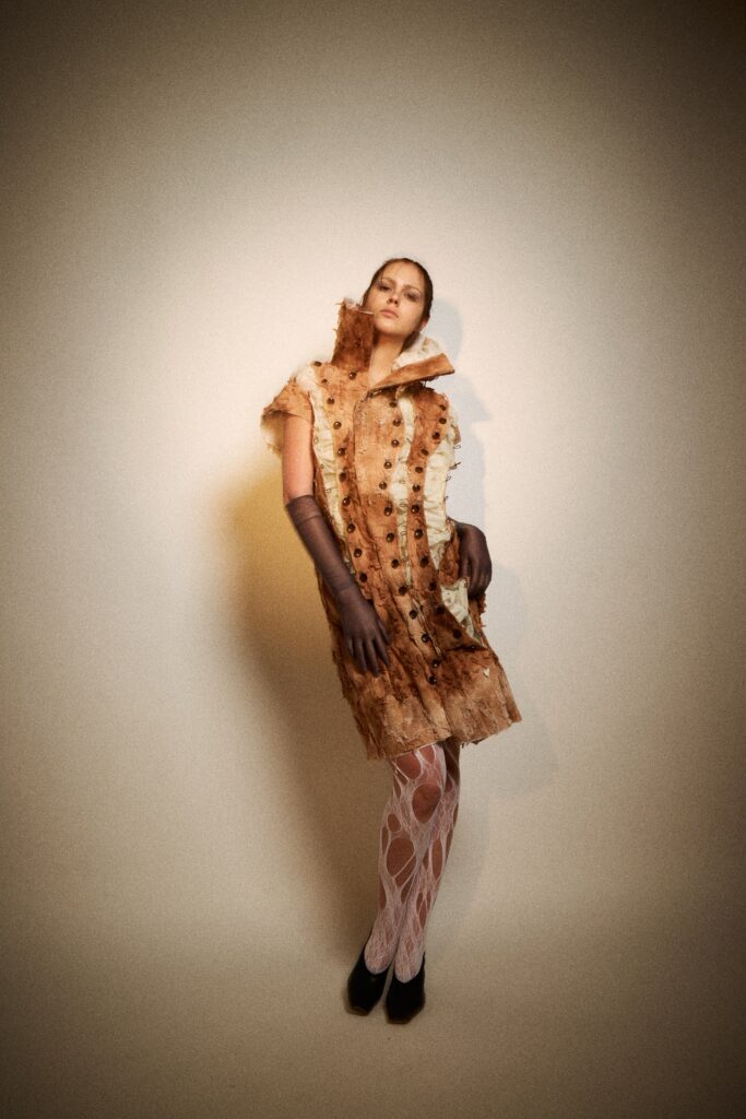 A model stands against a plain studio background wearing a pale rust-coloured textured dress with raw edges, exposed buttons, a high sculptural collar, grey gloves, white cut-out tights and black heels.
