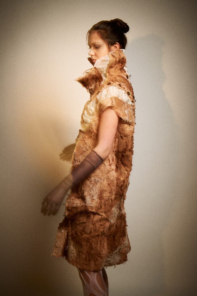 A model shown from the side wearing a pale rust-coloured textured dress with a high sculptural collar, raw edges, exposed buttons, grey gloves, white cut-out tights and black heels against a plain studio background.