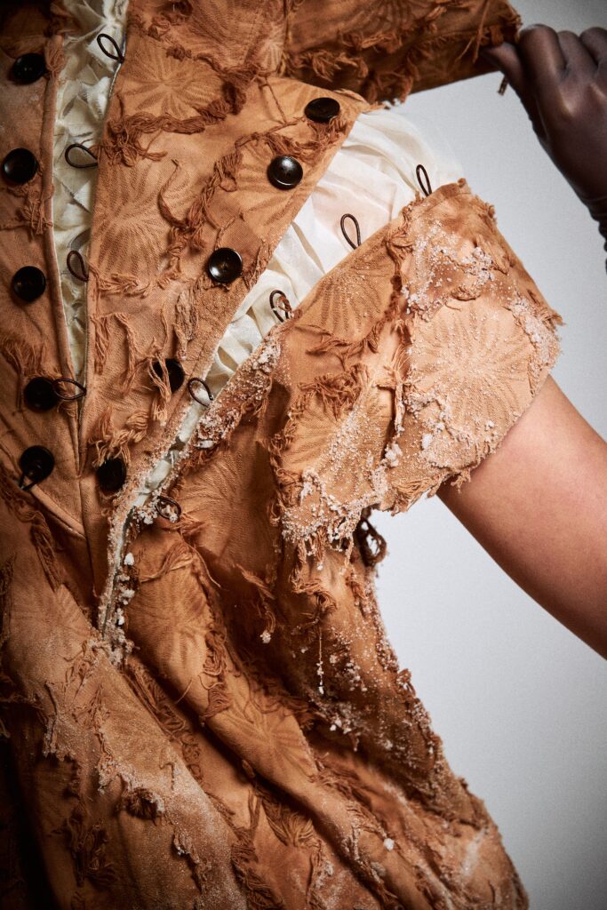 Close-up of a rust-coloured textured garment with raw fabric edges, black buttons, loop fastenings, white inner layers and small white alum crystals scattered across the surface.