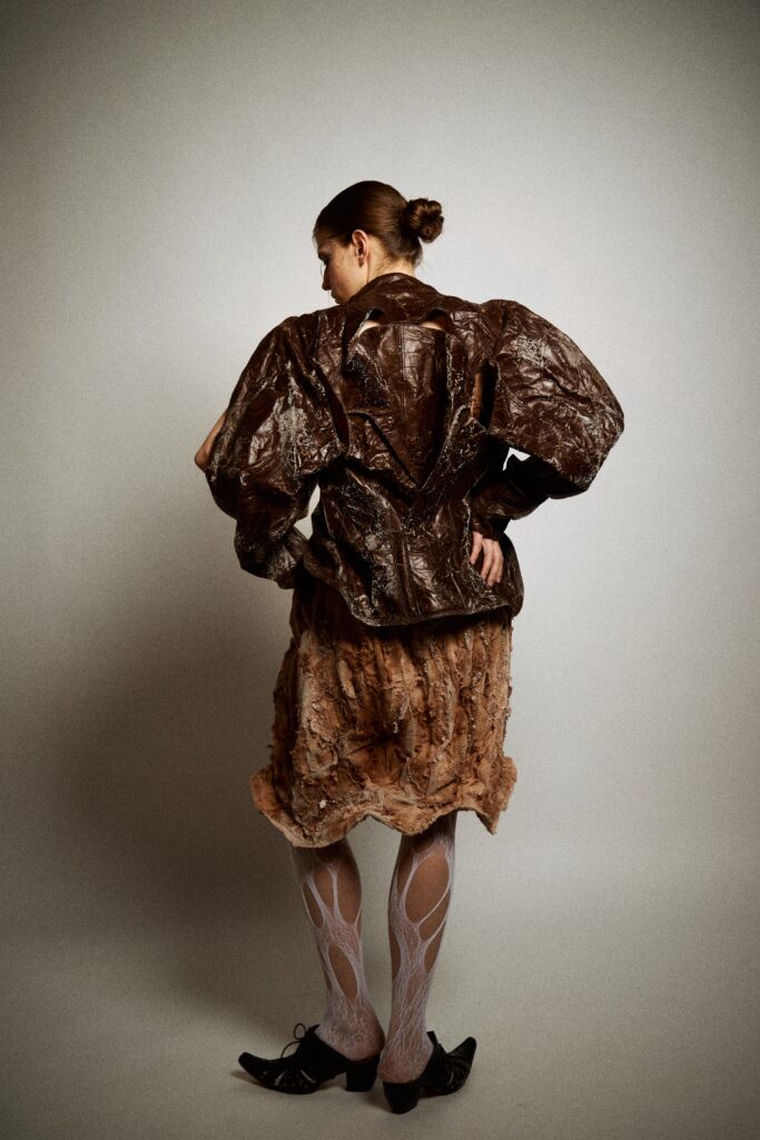 A model shown from the back wearing a dark brown sculptural jacket with oversized sleeves, cut-out openings, crystallised surface marks, a rust-coloured textured skirt, white cut-out tights and black heels against a plain studio background.