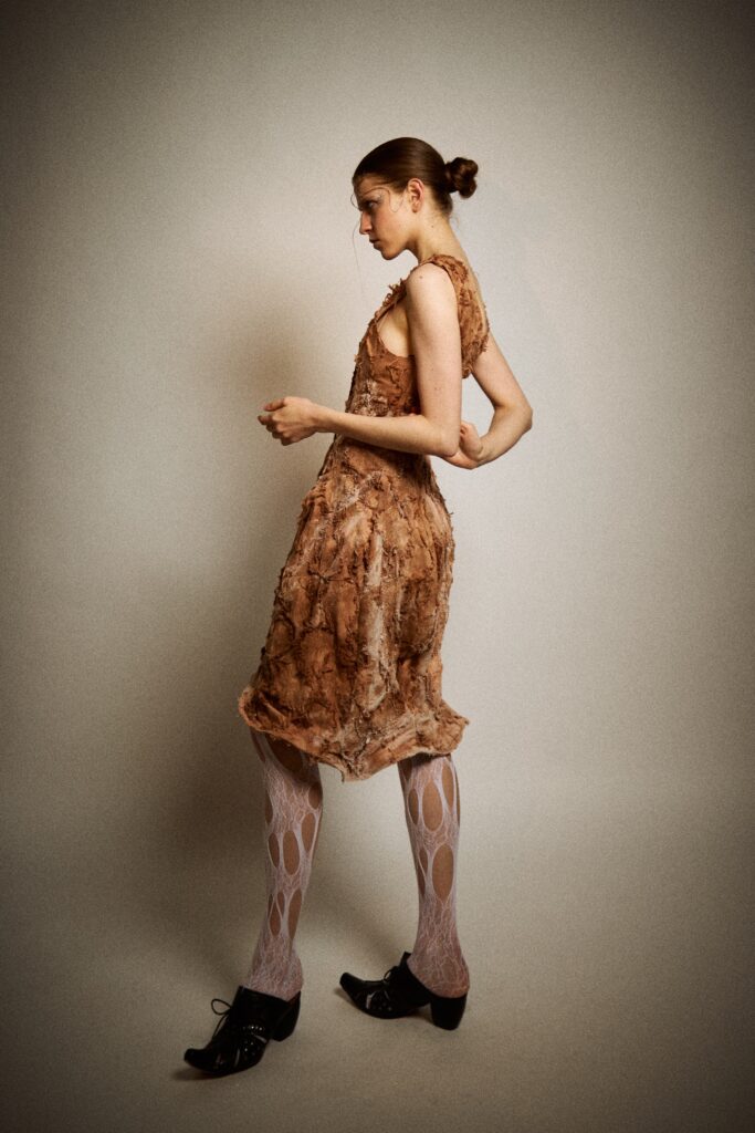 A model shown from the side wearing a rust-coloured textured skirt with raw edges, irregular surface details and subtle crystallised marks, paired with white cut-out tights and black heels against a plain studio background.