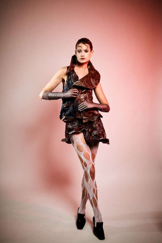 A model stands against a pink studio background wearing a dark brown and deep red textured fashion look with a sculptural collar, cracked leather-like surfaces, grey gloves, white cut-out tights and black heels.