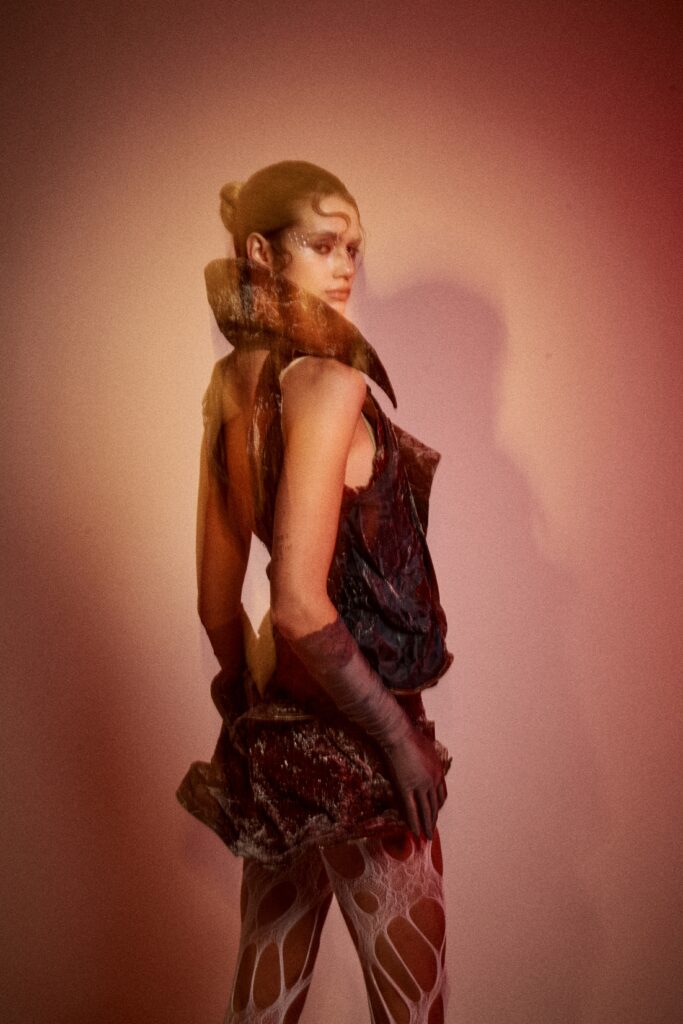 A model shown from the side against a pink studio background wearing a dark brown and deep red textured garment with an exposed back, sculptural collar, grey glove, white cut-out tights and black heels, captured with a blurred motion effect.