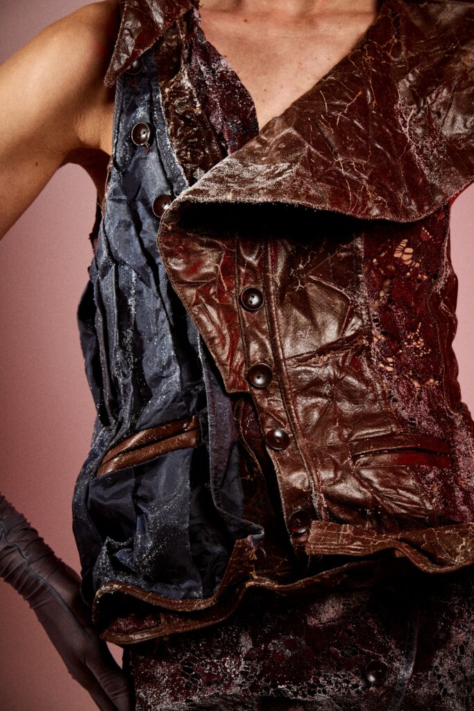 Close-up of a dark brown and deep red textured garment with cracked leather-like panels, black buttons, red lace, blue fabric layers and small white crystal deposits on the surface.