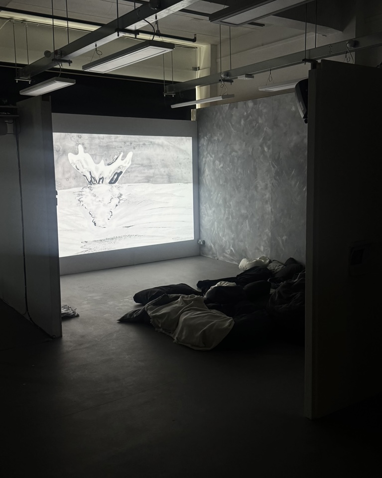 Image of projection in space. Walls of space are painted with a grey ‘lime wash’ effect and wall being projected onto is white. Space is furnished with various mis-match blankets, cushions and bean bags. The projection shows a splash of water.