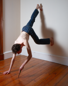 A young man balances between a wall and floor extending a knife toward the ground in an otherwise empty room.