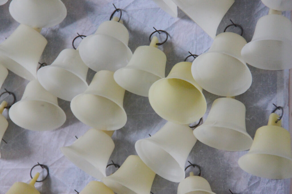 Close up installation shot of multiple yellow wax bells hanging from steel hoops and panel pins driven onto a board covered in translucent paper creased white.