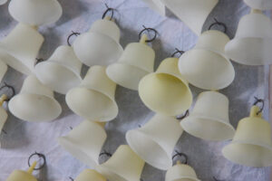 Close up installation shot of multiple yellow wax bells hanging from steel hoops and panel pins driven onto a board covered in translucent paper creased white.