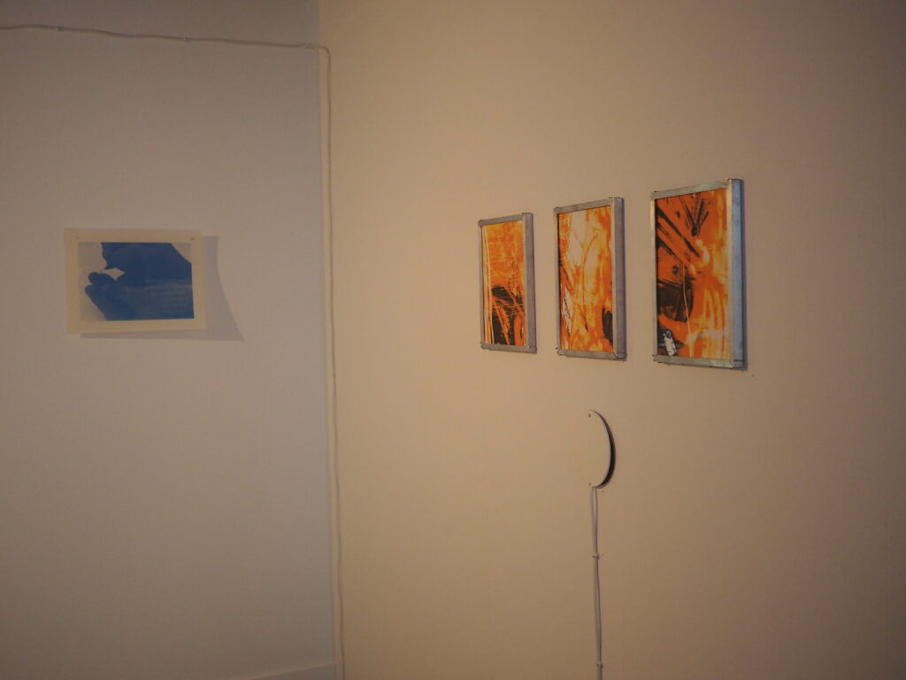 Blue of hands tattooing themselves on an adjacent wall to three orange prints framed in metal