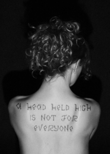 Black and white photograph of a person seen from behind, with handwritten text across their upper back reading “A head held high is not for everyone.”