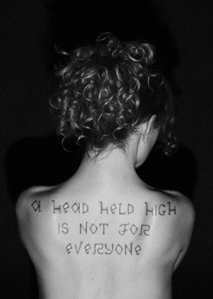Black and white photograph of a person seen from behind, with handwritten text across their upper back reading “A head held high is not for everyone.”
