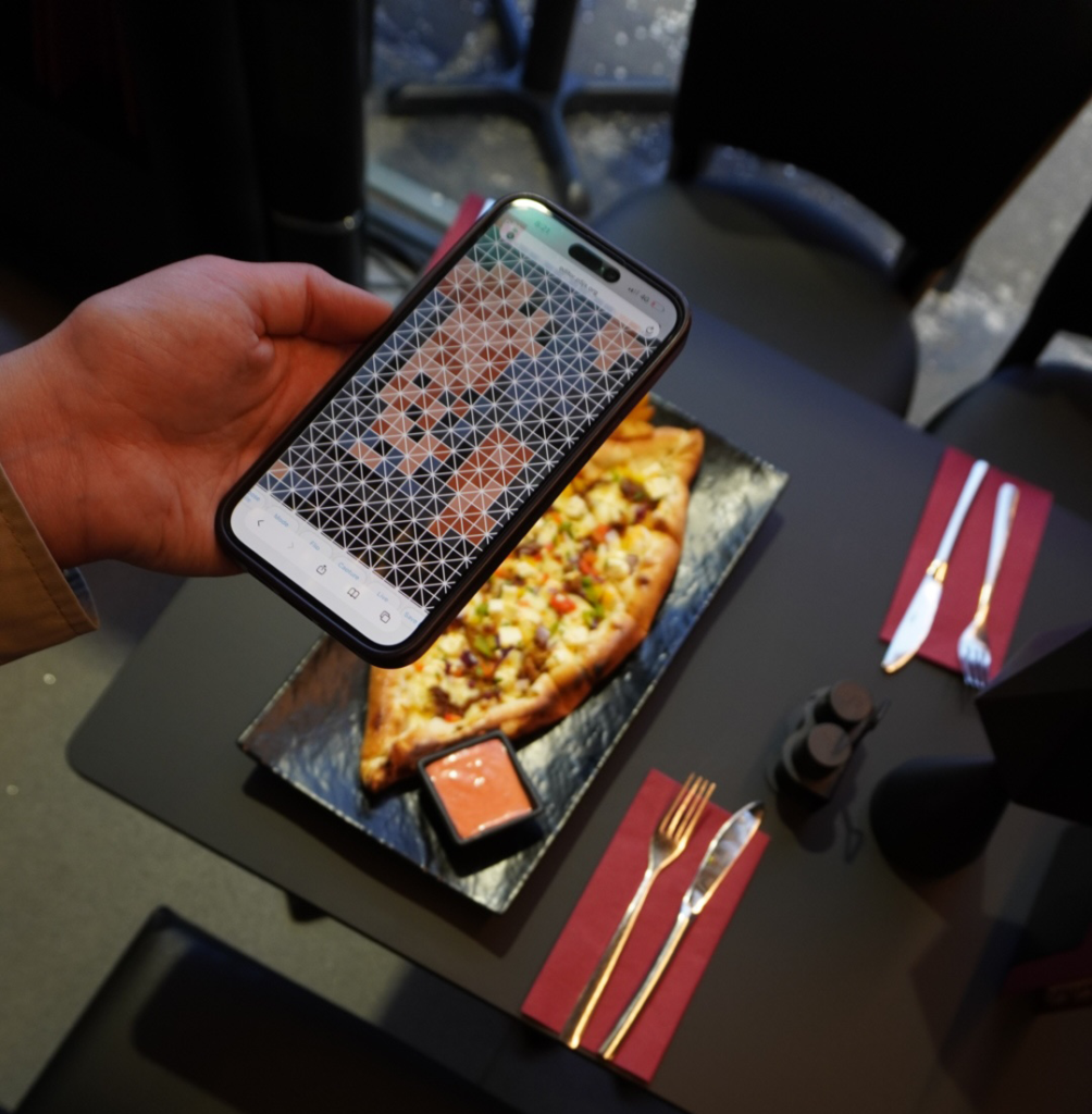A person holds a phone showing a mosaic-style image transformation above a table with a pizza and cutlery.