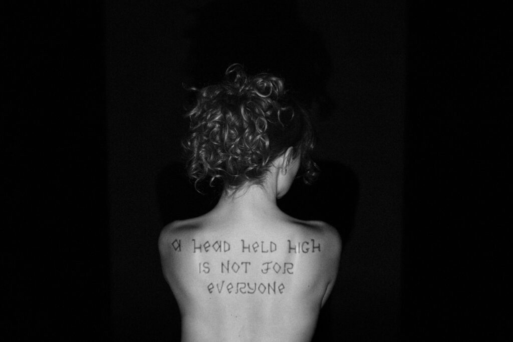 Black and white photograph of a person seen from behind against a dark background, with handwritten Latin text across their upper back reading 'A head held high is not for everyone'.