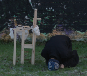 Performance documentation showing a figure kneeling beside a fragile sculptural structure in an outdoor environment.