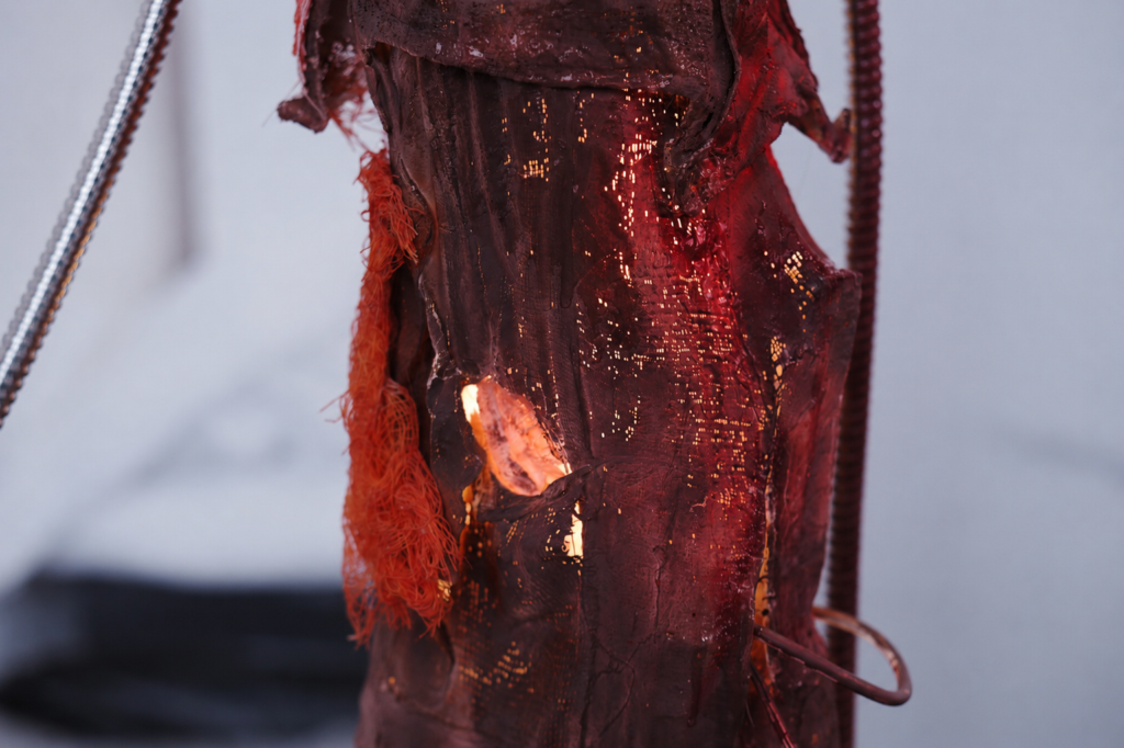 Close-up detail of a red illuminated pillar-like sculpture with layered silicone textures, exposed wire, and glowing internal light.